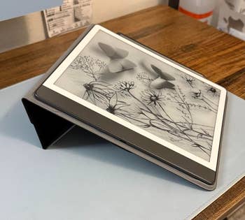 An e-reader successful  a achromatic  lawsuit  displays a grayscale botanical illustration with butterflies, resting connected  a woody  desk