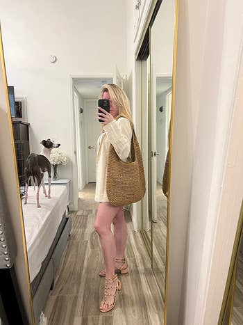 Woman in oversized blazer, holding a woven tote bag, takes a mirror selfie in a stylish room with a dog on the bed