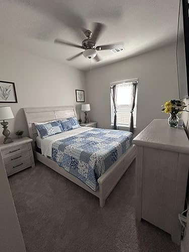 Cozy chamber  with a neatly made furniture  featuring a bluish  patterned quilt and matching pillows, a dresser with flowers, and framed partition  art