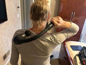 Person using a handheld massage tool on their back in a cozy room. The tool has a long handle and a massage node