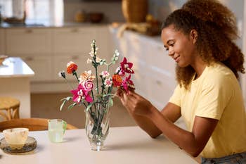 A person arranges a bouquet of colorful artificial flowers in a glass vase on a kitchen table