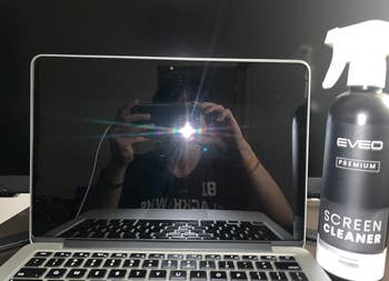 Person taking a photo with their phone, reflected on a laptop screen beside a screen cleaner bottle