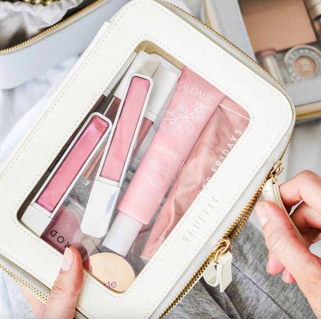 A person's hands hold a clear cosmetic bag filled with various beauty products, including lip gloss, a tube of Caudalie moisturizer, and other makeup items