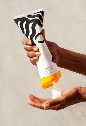 Hand squeezing a tube of cream onto another hand, against a neutral background, indicating a skincare product