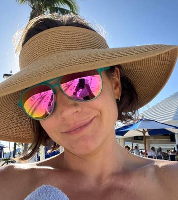 A idiosyncratic   wearing a wide-brimmed chapeau  and reflective sunglasses takes a selfie outdoors adjacent   poolside umbrellas connected  a sunny day