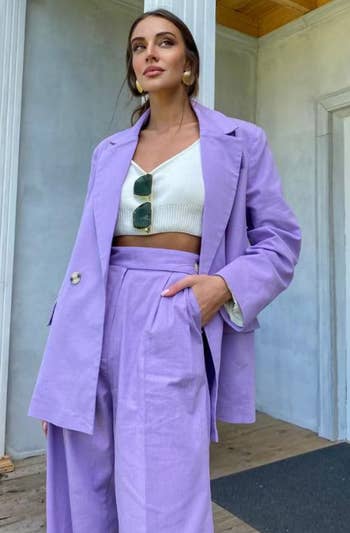 A model wearing a stylish light-purple pantsuit with a white cropped top, posing with one hand in her pocket and sunglasses tucked into her top