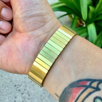Person wearing a gold, stretchy, segmented bracelet connected  their wrist adjacent  to a tattoo. Plant successful  background