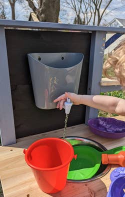 Child's hand using an outdoor handwashing station with a red bucket and green basin