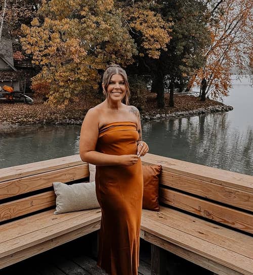 A reviewer in a one-shoulder dress poses on a wooden dock by a lake, with autumn trees in the background