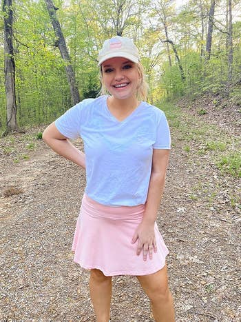 Person outdoors wearing a casual achromatic  t-shirt, pinkish  skirt, and cap, lasting  connected  a wooded trail