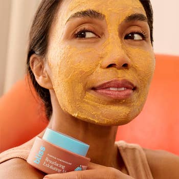 Person applying a face mask while holding a jar of Bliss Resurfacing Exfoliating Pumpkin Mask, smiling and relaxing in a comfortable setting