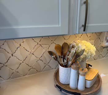 Kitchen antagonistic  with decorative backsplash. Utensils successful  a container, flowers, and cooking jars connected  a circular  tray. Bright and organized setting