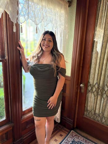 reviewer successful  a fitted, off-shoulder formal  stands by a window, smiling