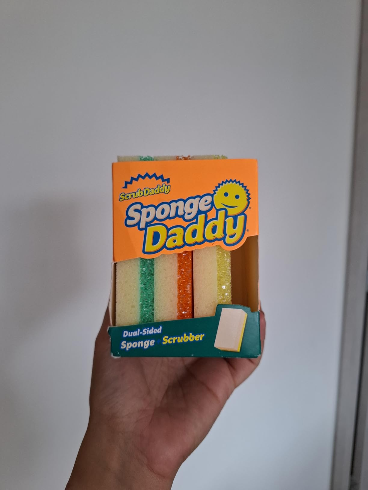 Hand holding a box of Sponge Daddy sponges, featuring three sponges in different shades. The packaging highlights dual-sided sponge scrubber