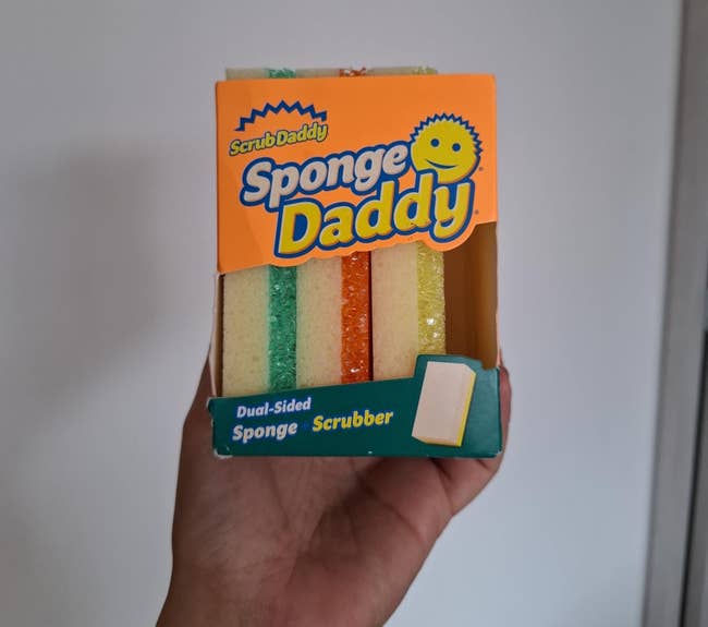 Hand holding a box of Sponge Daddy sponges, featuring three sponges in different shades. The packaging highlights dual-sided sponge scrubber