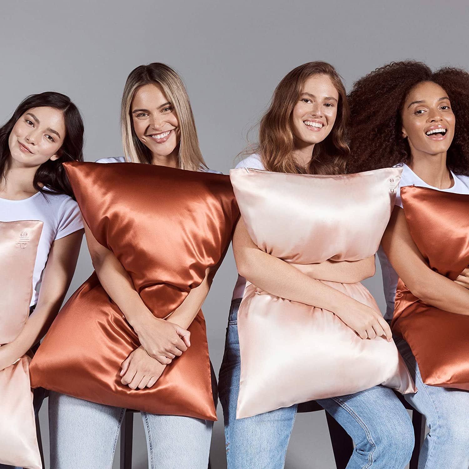four models holding pillows wrapped in pink and rust-colored silk pillowcases