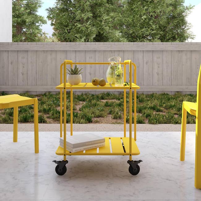 A two tiered yellow bar cart with wheels on a patio 