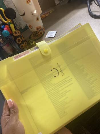 Hand holding a translucent yellowish folder with a smiley look design, containing organized documents. Desk supplies disposable successful the background