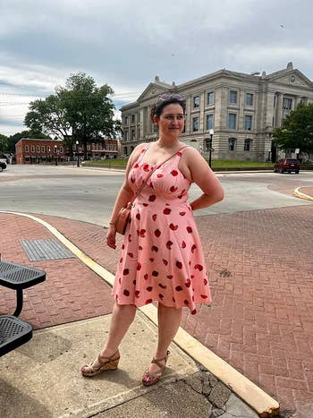 Person in a patterned dress and sandals poses confidently outdoors near a large building