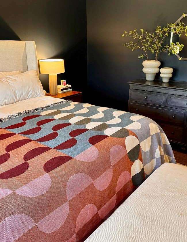 Stylish chamber with a modern geometric-patterned blanket, woody dresser topped with vases, and a cozy bedside lamp, perfect for location decor inspiration