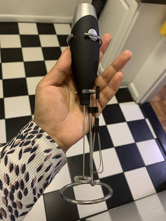 Reviewer holding black and silver milk frother attached to a silver stand and mini black and silver whisk