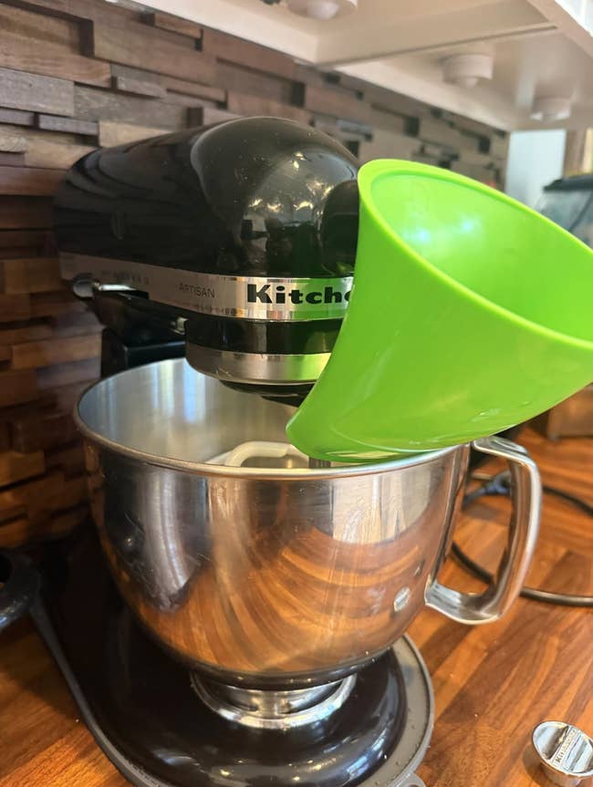 Stand mixer with a stainless alloy vessel and a greenish pouring shield, placed connected a room counter