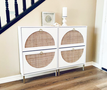 Two white slim entryway storage holders with rattan shaped in half moons small gold handles to open oup the two compartments on each holder 
