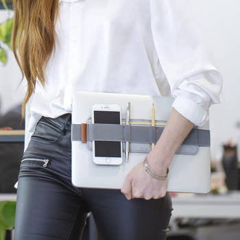 Model carrying their laptop with the elastic laptop organizer holding a phone, notebook, pen, and mouse