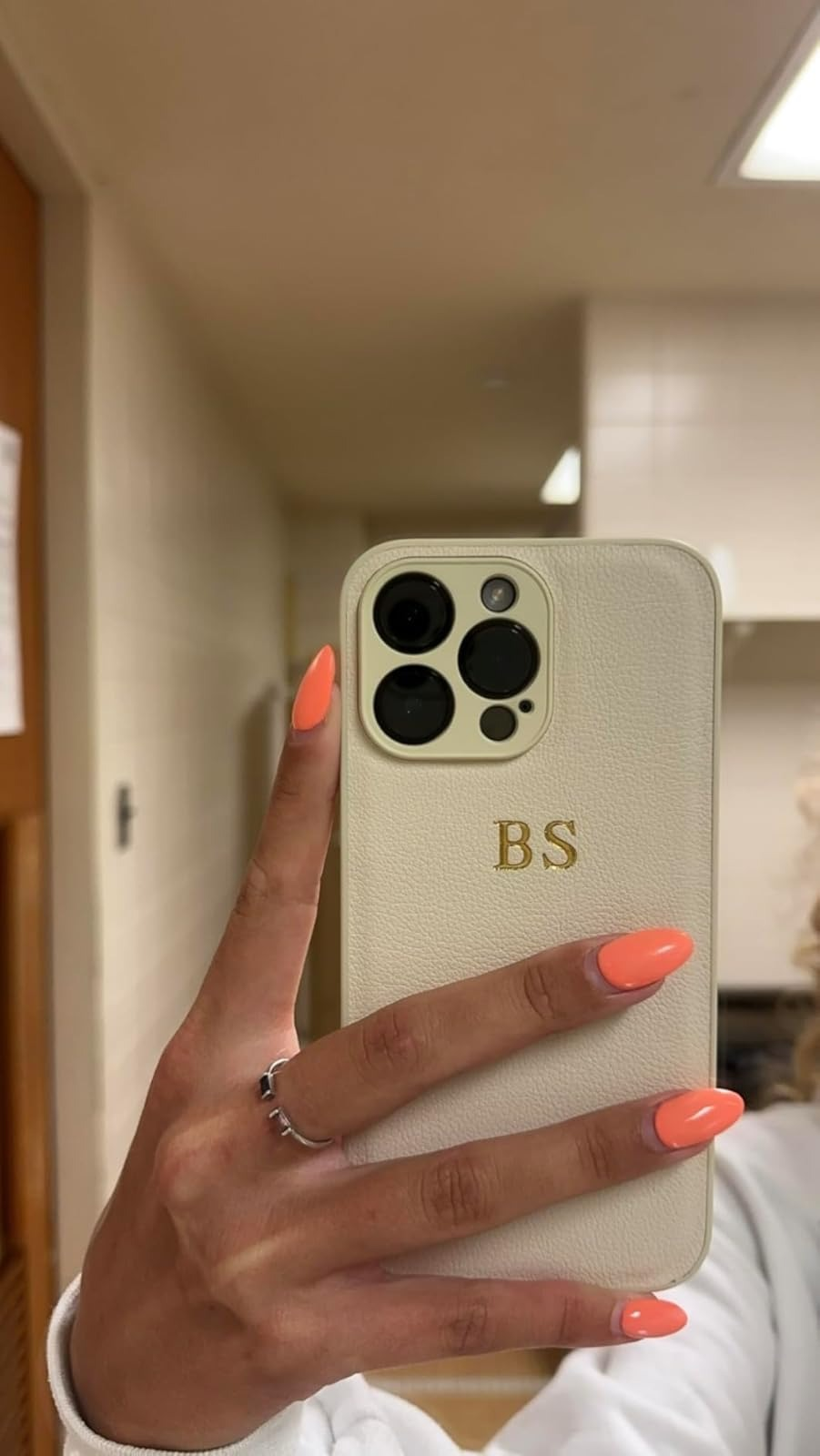 A hand with bright orange manicured nails holds up an iPhone with a personalized white case, featuring the initials 
