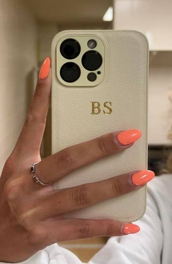 A hand with bright orange manicured nails holds up an iPhone with a personalized white case, featuring the initials