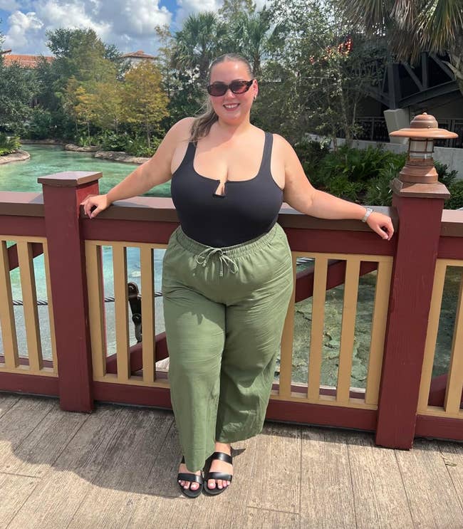 Person smiling outdoors, posing by a woody  railing. She is wearing a sleeveless achromatic  top, greenish  wide-leg pants, sunglasses, and sandals