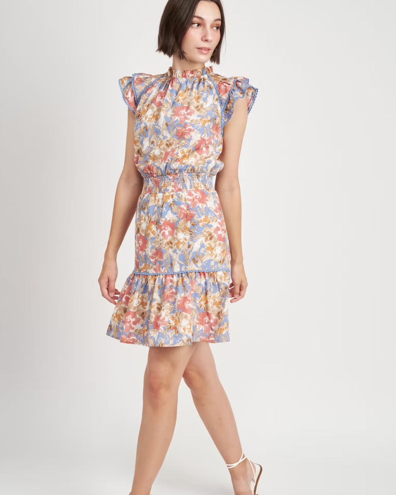 Model in a blue dress with green and pink watercolor pattern that has a high neck, ruffled sleeves, and cinches at the waist 