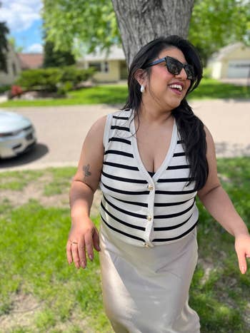 reviewer outdoors, smiling successful striped sleeveless apical and airy skirt, wearing sunglasses and earrings