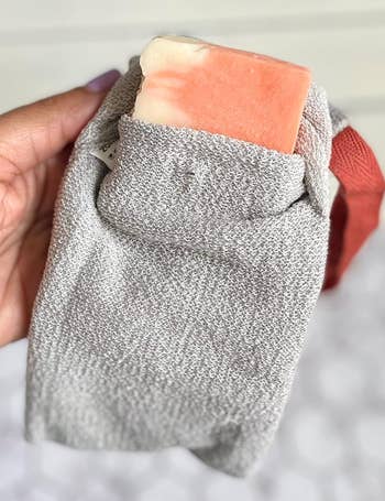 A hand holds a textured gray exfoliating washcloth pocket with a bar of soap partially inserted at the top