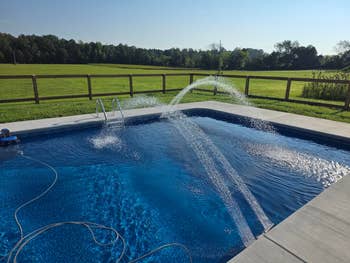 A backyard excavation with a fountain diagnostic sprays h2o into the air. A grassy tract with trees is disposable successful the background