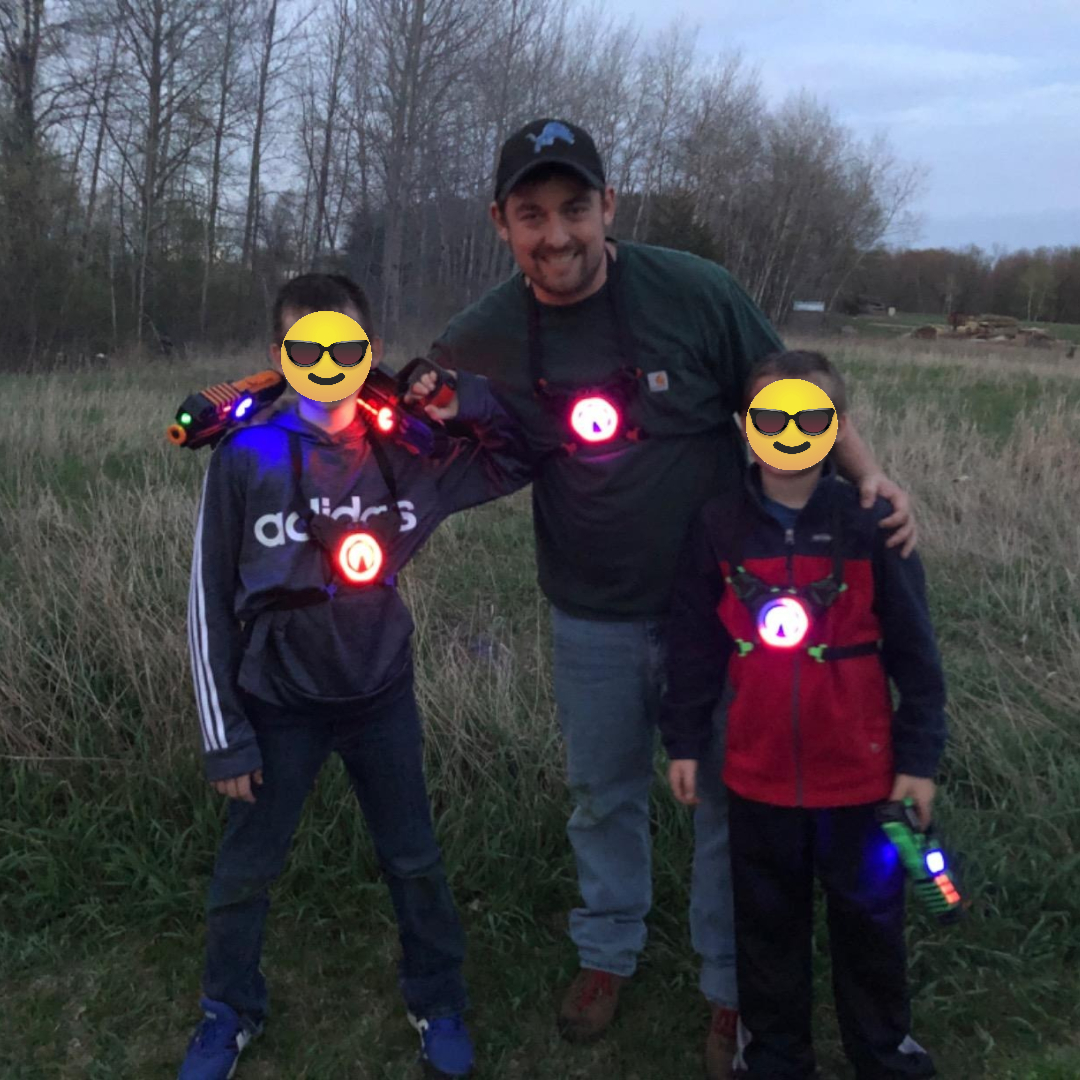 Three people smiling outdoors, wearing laser tag gear with glowing targets. Two children stand with a man in casual clothes, holding laser guns