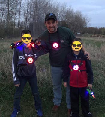 Three people smiling outdoors, wearing laser tag gear with glowing targets. Two children stand with a man in casual clothes, holding laser guns