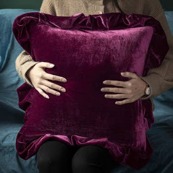 Person holding a large, ruffled velvet pillow, sitting. Sweater and casual attire visible