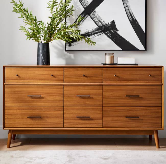 Wooden dresser with six drawers, sleek handles, and angled legs in a room with a plant and wall art