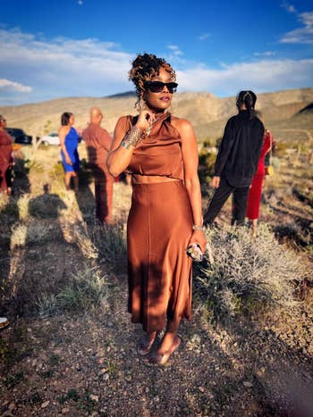 A woman stands confidently in a desert landscape, wearing an elegant sleeveless dress and sunglasses, holding a small clutch. Others are visible in the background