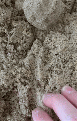A close-up of a hand playing with a ball of kinetic sand, smoothing and shaping the sand texturally