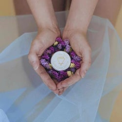 Hands holding purple flowers and clitoral balm with earth sign sigil
