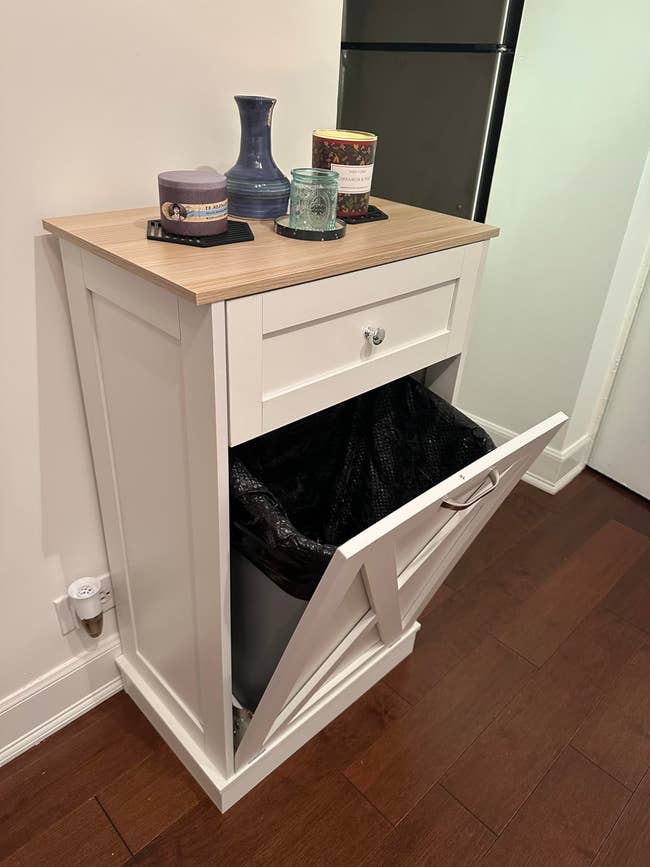 Small white kitchen cabinet with a wooden top holding candles and a vase, featuring a pull-out trash bin on the lower half