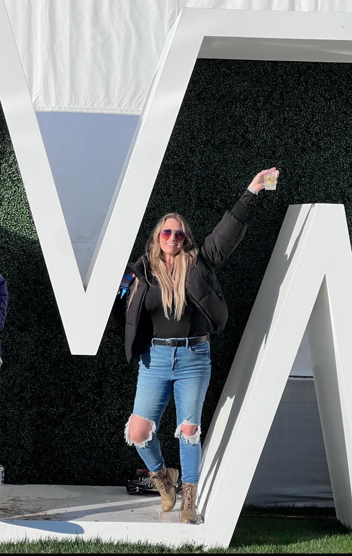 Person posing casually with ripped jeans and brownish  boots, holding a drink. They are lasting  playfully against a ample  achromatic  missive  structure