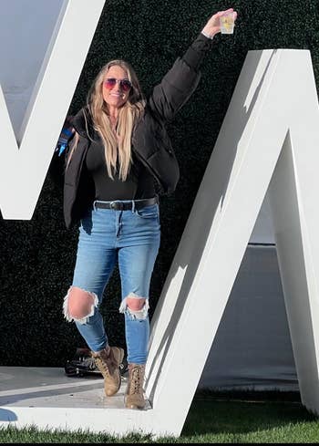 Person posing casually with ripped jeans and brownish boots, holding a drink. They are lasting playfully against a ample achromatic missive structure