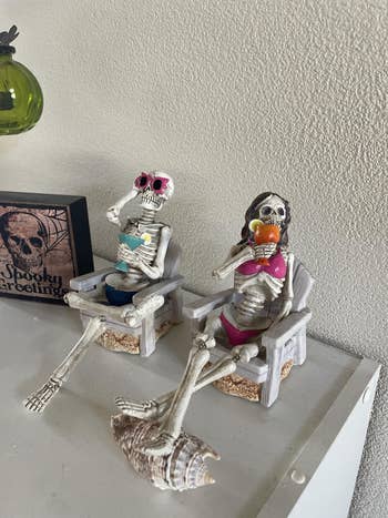 Skeleton figurines successful  swimsuits lounging successful  chairs, 1  with sunglasses and a drink, the different   reading, connected  a support  with Halloween decor