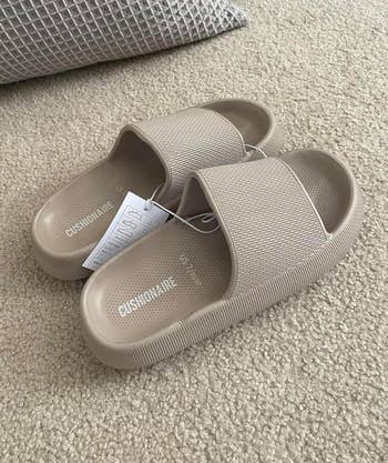 A pair of CushioNaire slide sandals are on a carpet with a tag attached. A cushiony mat is partially visible in the background