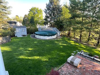 Backyard with covered circular  pool, adjacent shed, and patio area. Lush greenish  tract   surrounded by trees and garden