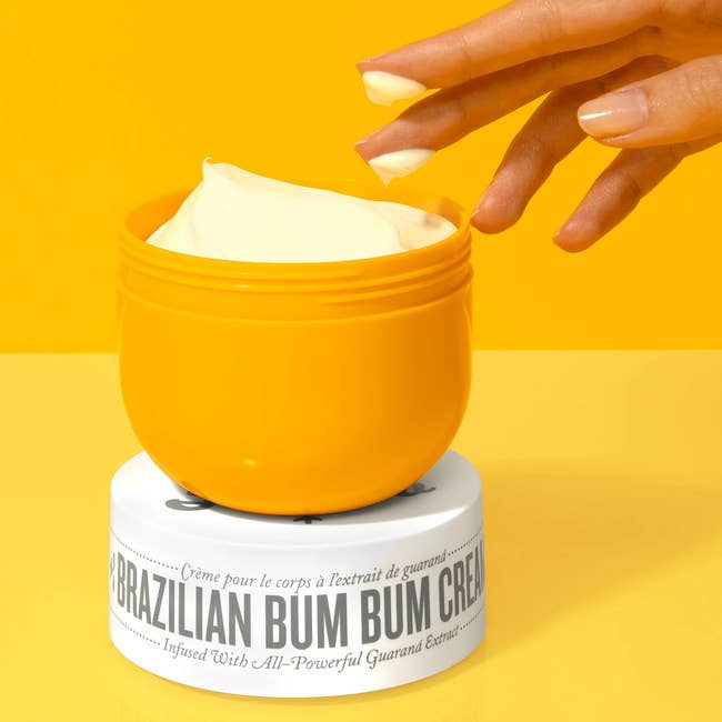 a hand with product on two fingers from the open yellow tub of body cream