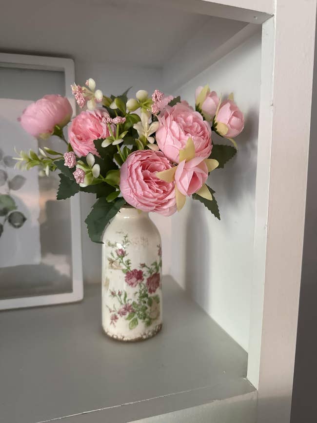 Floral statement with pinkish peonies and greenish leaves successful a decorative achromatic vase connected a shelf